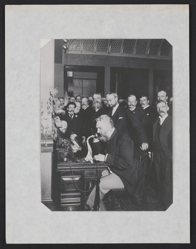 Alexander Graham Bell seated at a carved desk with a receiver to his ear, speaking into an upright desk telephone, watched by about a dozen men in dark suits