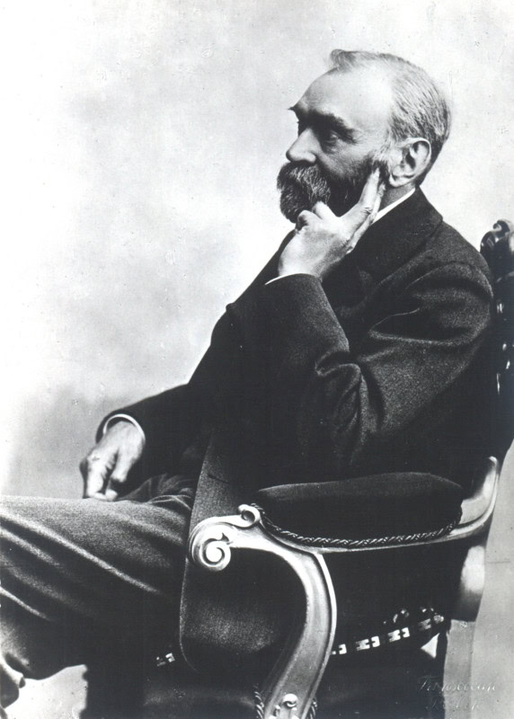 Studio photograph of Alfred Nobel in profile, seated in a carved armchair in a dark suit with one hand at his bearded chin