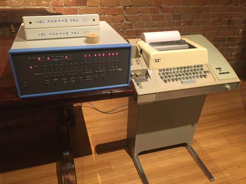 A MITS Altair 8800 computer with a blue-grey front panel of toggle switches and lit red indicator lights, standing beside a cream Teletype Model 33