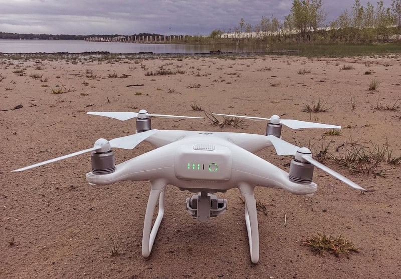 A white DJI Phantom 4 quadcopter drone standing on gravel with its four rotors and camera gimbal visible