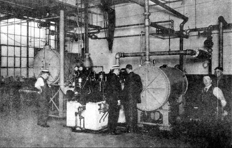 Black-and-white photograph of an early air-conditioning machine room: two large horizontal cylindrical vessels, an engine-driven compressor and heavy overhead pipework, with five men in shirtsleeves and hats among the plant
