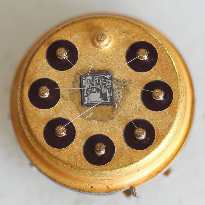 An opened round gold metal-can integrated circuit seen from above, the square silicon die at the centre with fine gold bond wires fanning out to eight pins