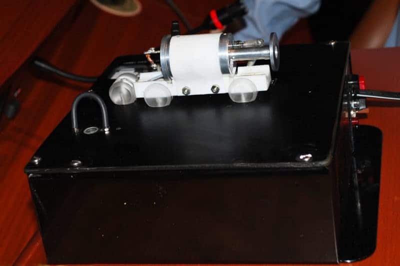 An early ruby laser: a small white cylindrical head with metal end mirror and flashlamp fittings, sitting on a black power-supply box on a wooden table
