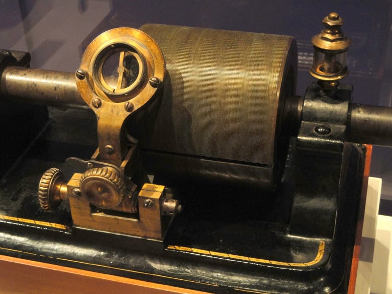 An Edison tin-foil phonograph: a grooved brass cylinder on a crank shaft with a gear and a mica diaphragm arm, mounted on a black wooden base
