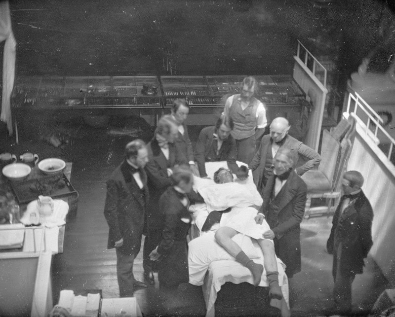 Daguerreotype looking down into an operating theatre, with seven frock-coated men gathered around an unconscious draped patient on a table and glass instrument cases behind
