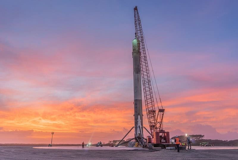 A Falcon 9 first stage standing upright on its landing legs at Landing Zone 1 at sunset, with a crane alongside