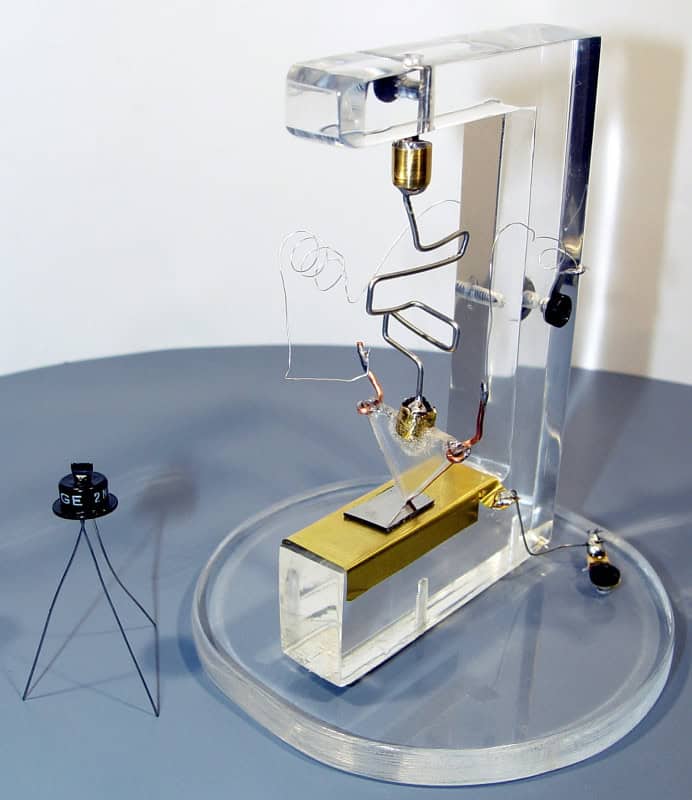 Replica of the 1947 Bell Labs point-contact transistor: a triangular plastic wedge with gold-foil contacts pressed onto a germanium slab, wire leads, on a clear acrylic stand, with a small modern transistor beside it for scale