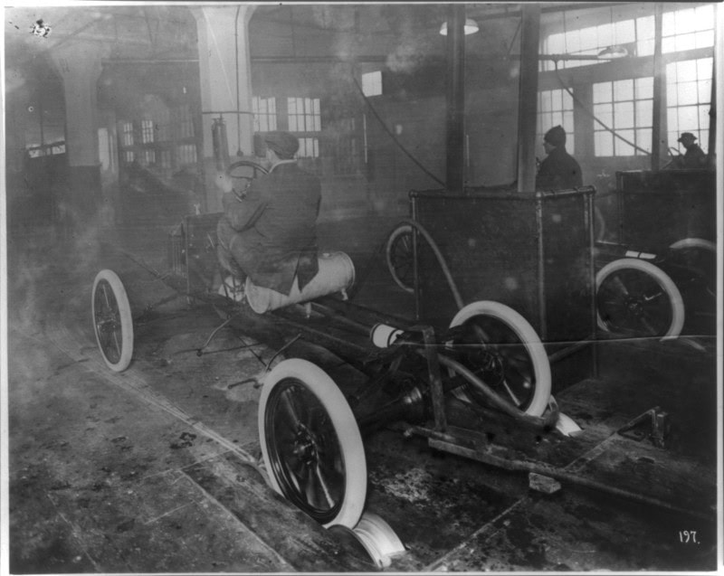 A bare Model T chassis on spoked wheels on a factory floor, a workman in cap and overalls astride it fitting parts, with part-built bodies and other workers in the windowed hall behind
