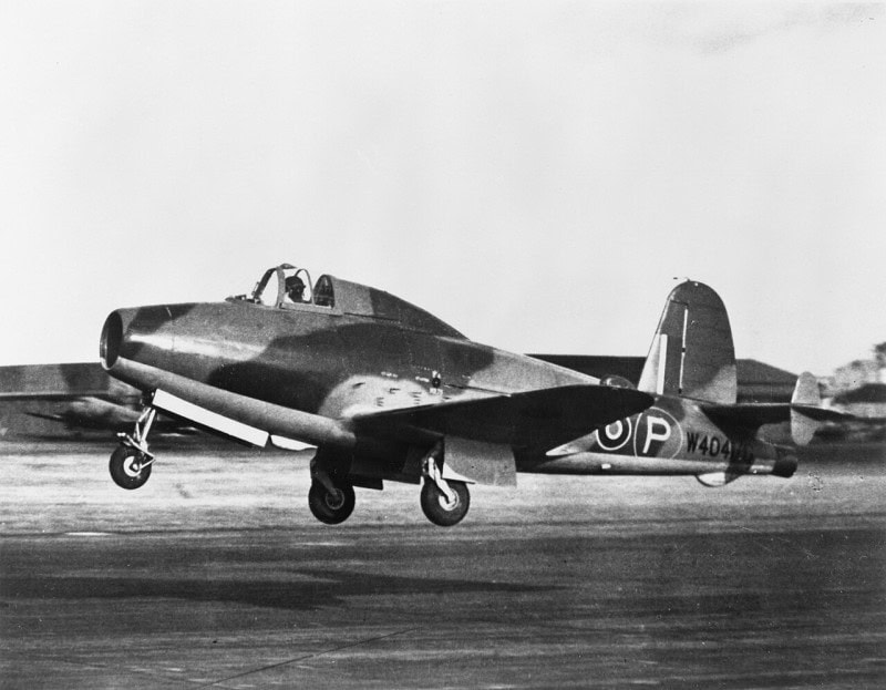 The Gloster E.28/39 jet aircraft just lifting off a runway, camouflaged, with an RAF roundel and the serial W4041 on the fuselage and hangars behind