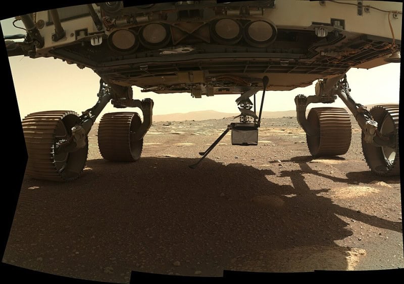The Ingenuity helicopter stowed beneath the belly of the Perseverance rover, photographed against the Martian ground before deployment