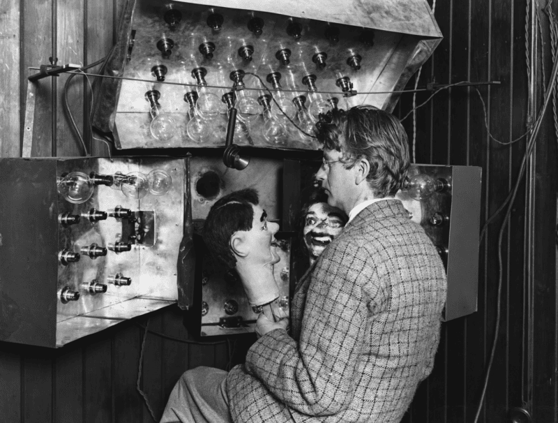 John Logie Baird in a tweed jacket holding a ventriloquist's dummy head up to his televisor apparatus, a large metal frame studded with dozens of bare light bulbs
