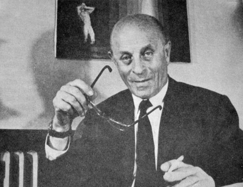Black-and-white portrait of an elderly, bald László Bíró in a dark suit, holding his glasses, with framed pictures on the wall behind