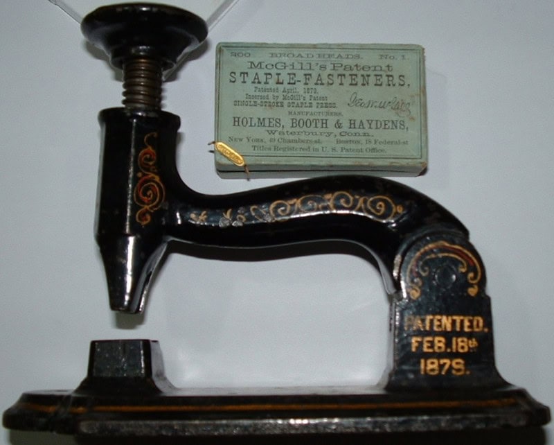 A black cast-iron McGill Single-Stroke Staple Press with gold pinstriping, its base lettered "Patented Feb. 18th 1879", beside its original green box