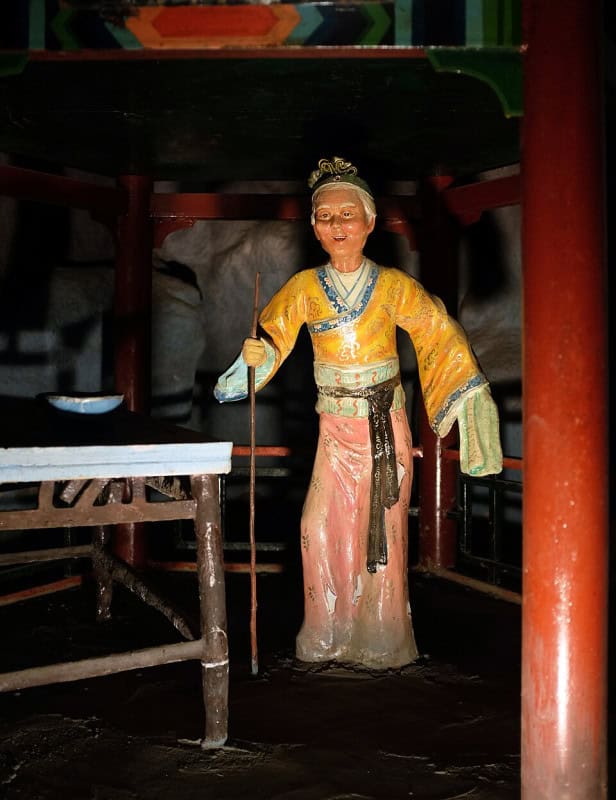 Painted statue of Meng Po as a grey-haired old woman in a yellow jacket and pink skirt, leaning on a long thin staff, with a white bowl on a table beside her at the Pavilion of Forgetfulness, Haw Par Villa