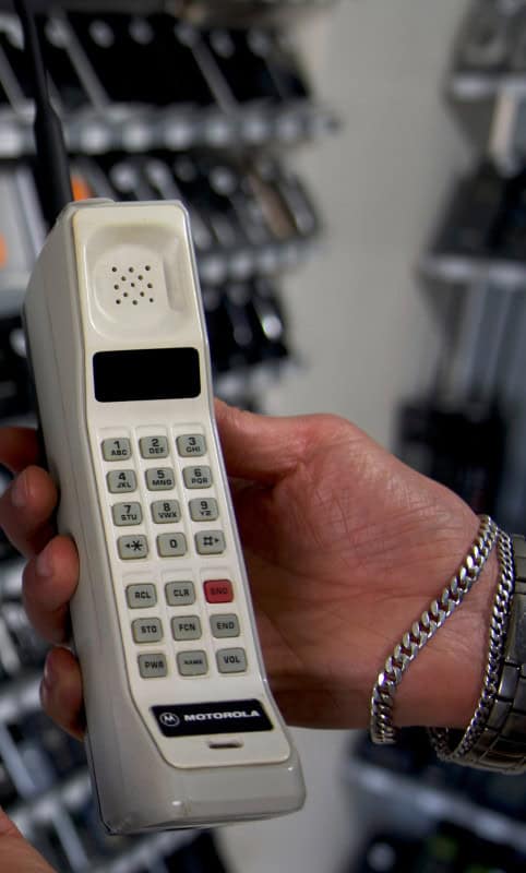 A hand holding a cream Motorola DynaTAC mobile phone, showing its earpiece grille, dark display and grey keypad, with a blurred rack of modern phones behind