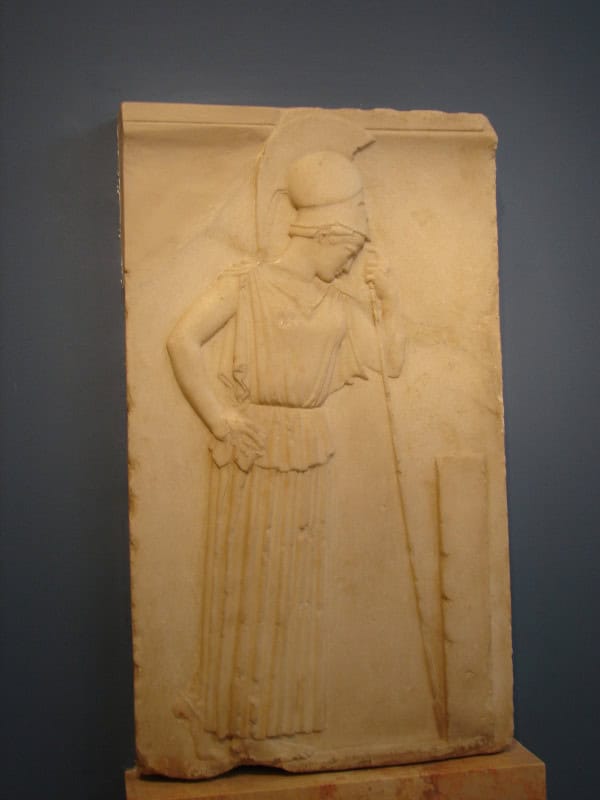 Low marble relief of a helmeted Athena leaning on her spear with her head bowed towards a stone pillar