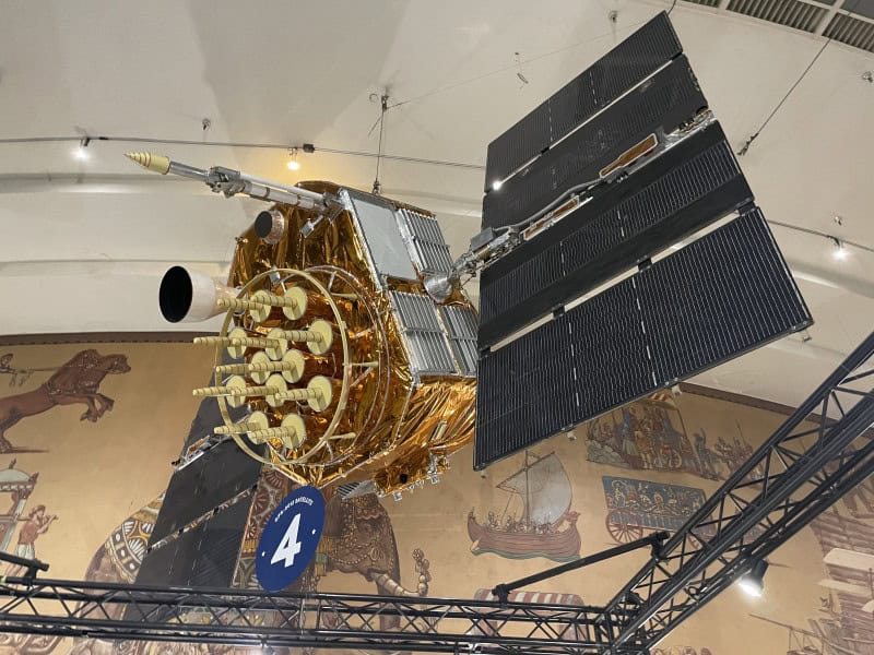 A NAVSTAR GPS satellite suspended from a museum ceiling, wrapped in gold foil with two black solar wings and a circular array of cream helical antennas facing the camera