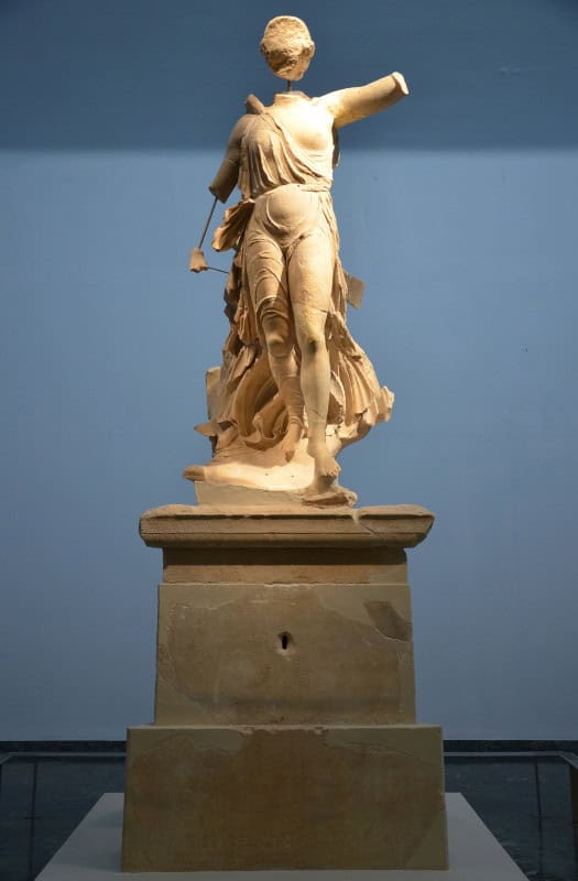 The weathered Olympia marble of the Nike of Paionios on its pedestal, face and arms lost, drapery pressed back against her legs