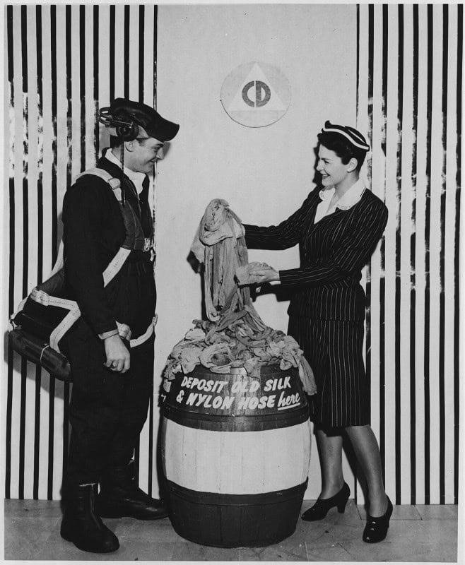 A woman in a pinstripe uniform dropping stockings into a wooden barrel stencilled "Deposit Old Silk & Nylon Hose here", with a man in a parachute harness standing opposite