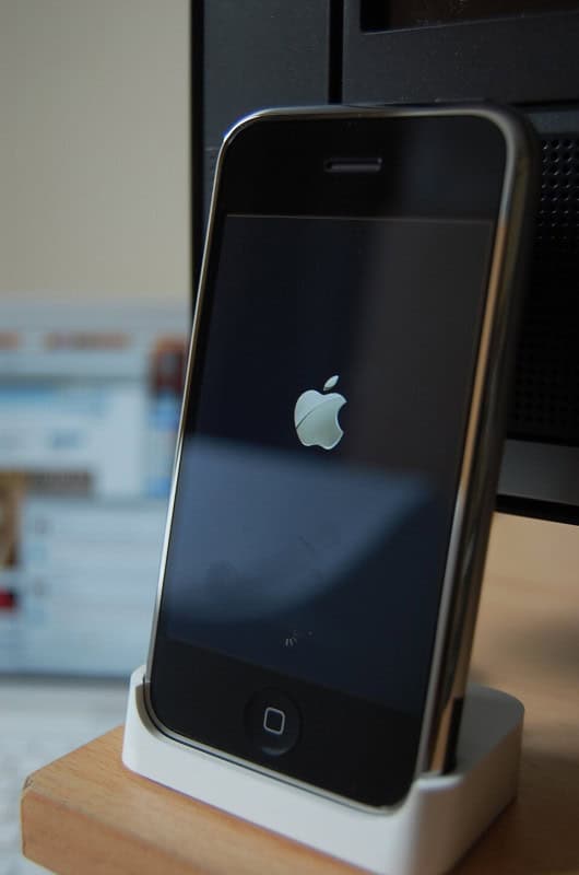 An original 2007 iPhone standing in its white dock, its screen showing the Apple logo