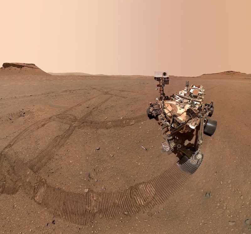 Self-portrait of the Perseverance rover standing on reddish Martian ground, with its wheel tracks curving away across the surface behind it