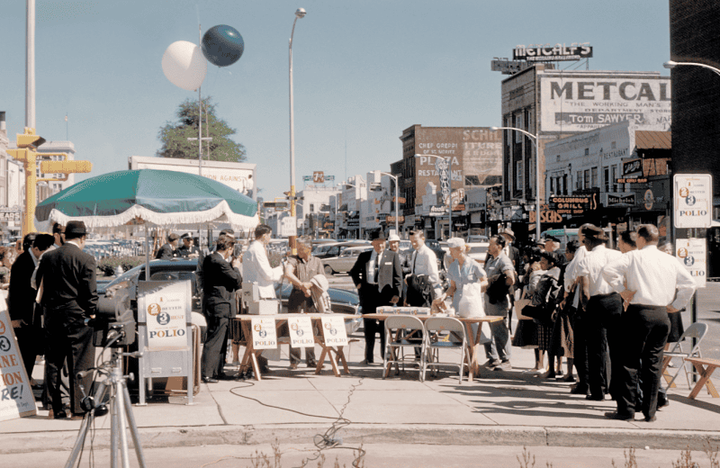 A street-corner polio vaccination clinic: a crowd at trestle tables under a fringed umbrella, placards reading "1 2 3 Better Best Polio", period cars and a department store behind
