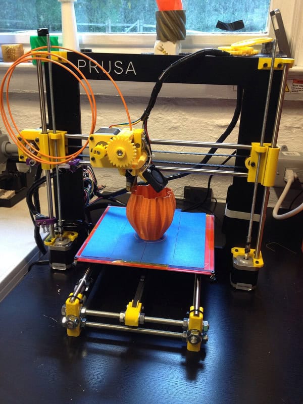A Prusa i3 RepRap desktop 3D printer mid-print, building an orange object on a blue print bed
