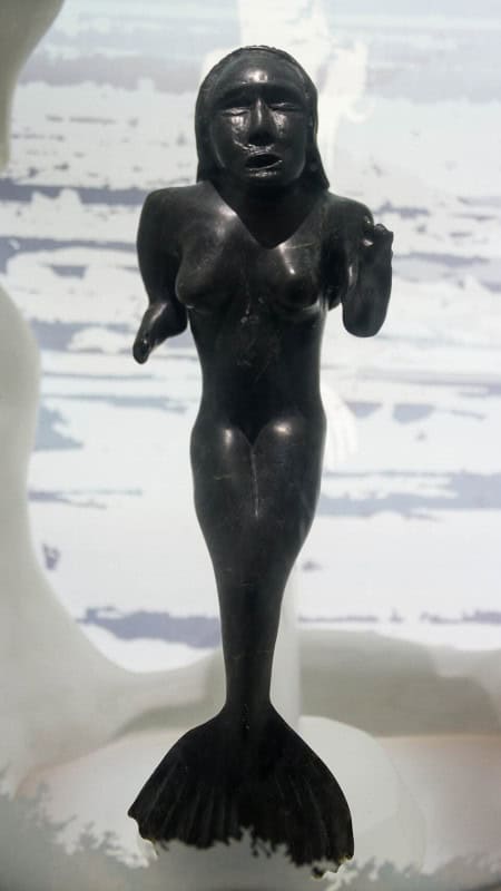 Dark stone sculpture of Sedna standing on snow, a female figure whose lower body ends in a sea-mammal tail