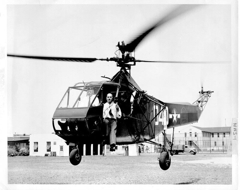 A single-rotor helicopter hovering a few feet above grass with its rotors blurred, one man at the controls in the glazed cabin and a second standing on the step