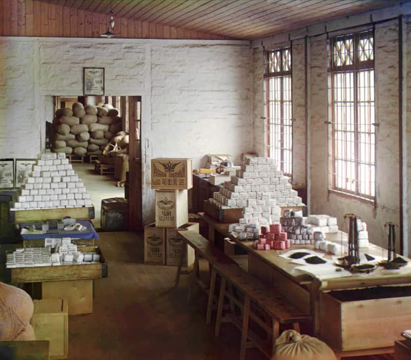 Early colour photograph of a whitewashed tea packing room, with tall pyramids of small white paper tea packets stacked on trestle tables and balance scales and trays of loose black tea in the foreground