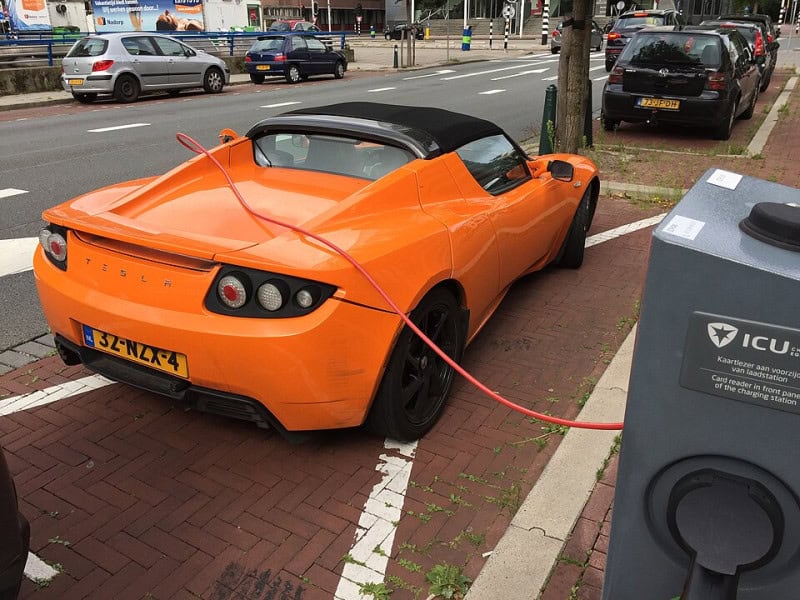 An orange Tesla Roadster parked at a kerbside charging point with its charging cable connected