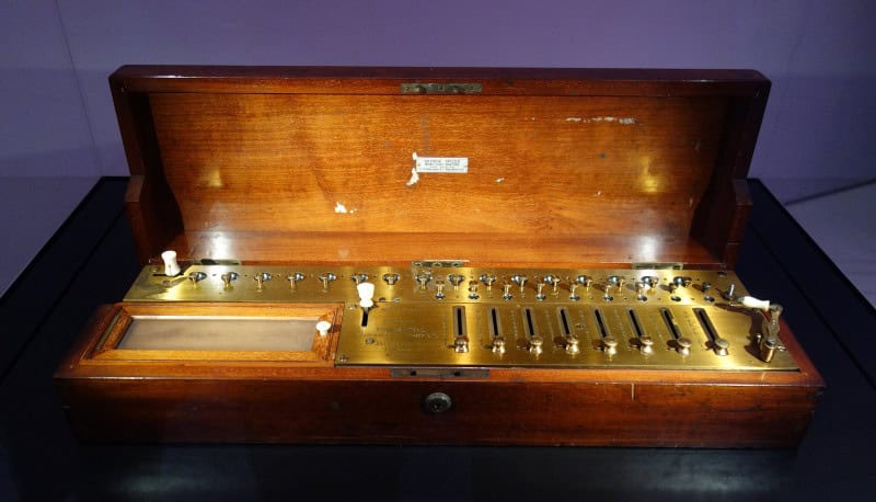 An arithmometer in an open mahogany case, with a long engraved brass faceplate, a row of small knobs, ivory-tipped sliding levers and result windows