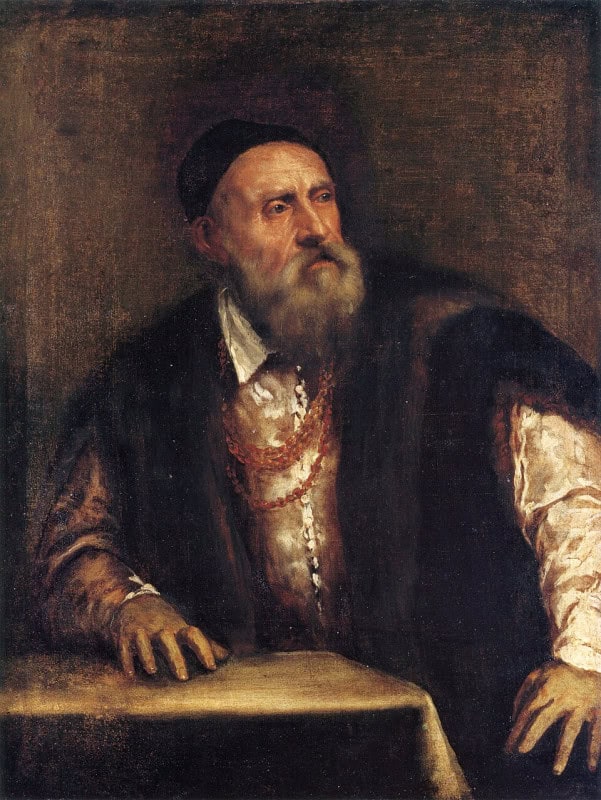 Elderly man with a long white beard in a dark cap and fur-trimmed robe over a white shirt, a gold chain across his chest, both hands resting on a table as he turns his head away to one side