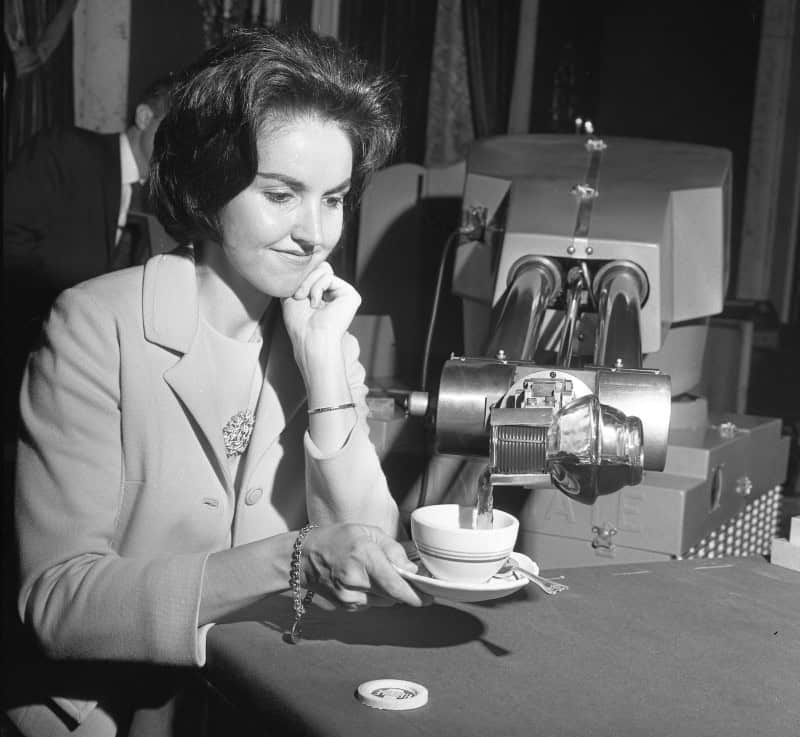 A woman resting her chin on her hand, watching a large chrome Unimate robot arm pour coffee into a cup