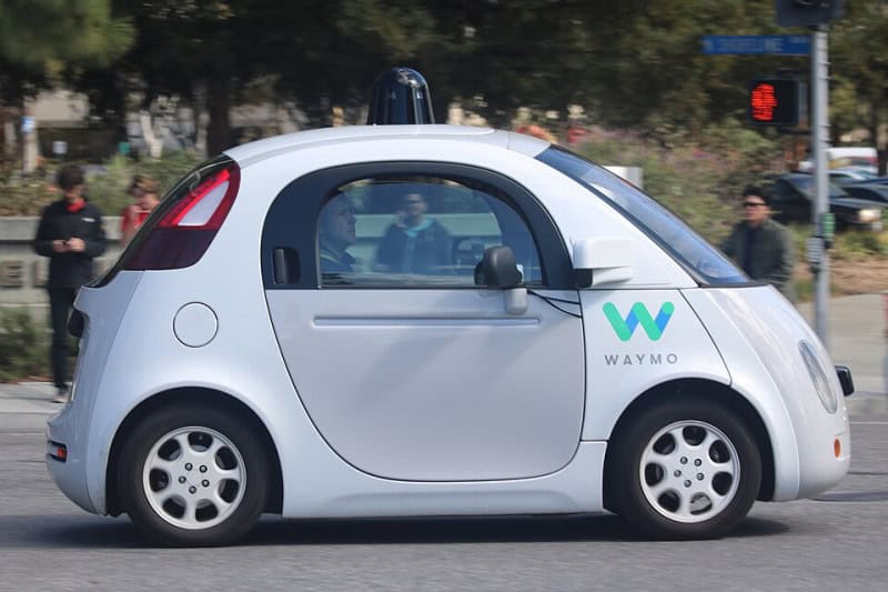 A white Waymo self-driving car with a roof-mounted sensor unit, photographed on a city street