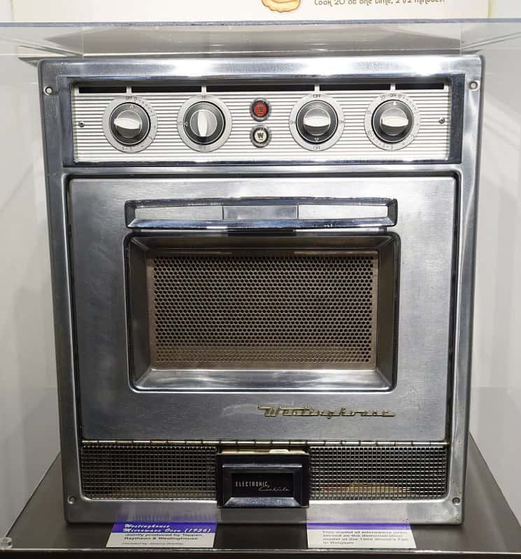 A 1956 Westinghouse microwave oven with a chrome front, four large dials across the top and a mesh-screened window, displayed in a glass museum case
