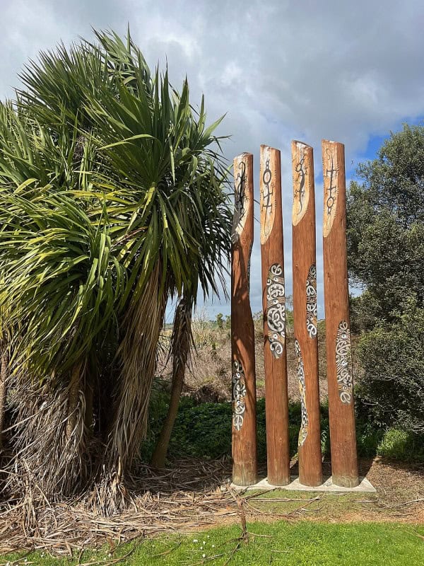 A row of carved wooden pou standing in grassland beside a cabbage tree at Lake Ngaroto