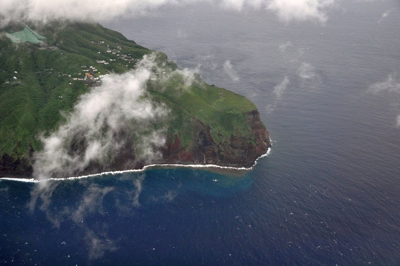Aerial photograph of Aogashima showing sheer dark cliffs, green slopes and a small village on the headland with cloud drifting over