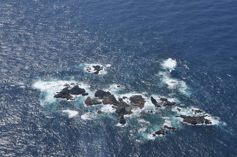 Aerial photograph of the Bayonnaise Rocks, a cluster of dark rocks ringed by white surf in deep blue open ocean