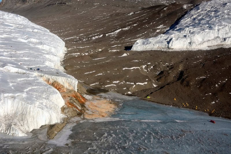 A rust-red stain spreading down the white ice cliff of the Taylor Glacier onto the frozen lake below, seen from above with a small field camp nearby