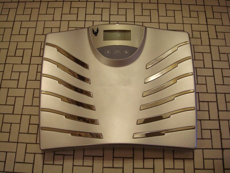 A digital bathroom scale standing on a tiled floor with a blank display