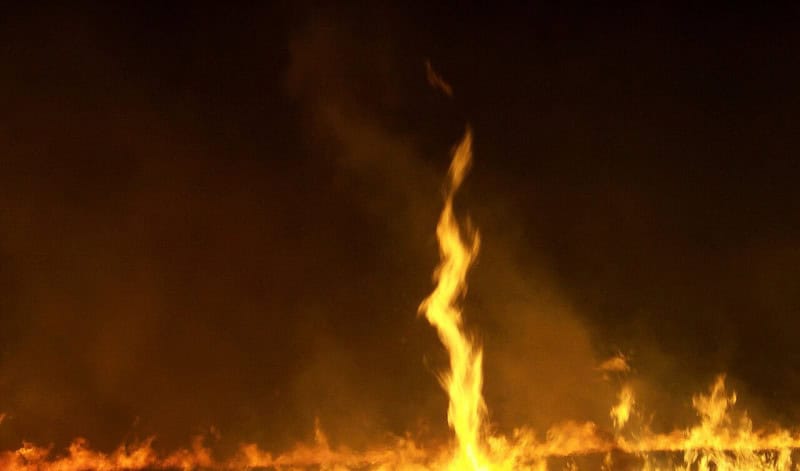 A rotating column of flame rising out of a line of burning grass at night