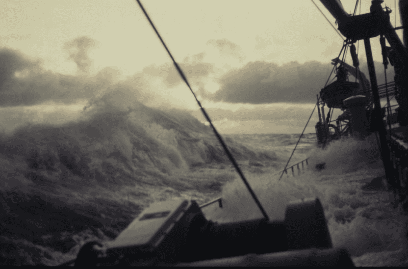 Colour photograph from a ship in heavy seas, with a large grey wave breaking over the deck and hatch covers