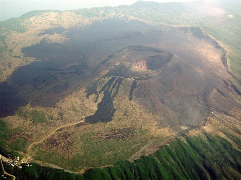 Aerial view of the bare brown summit crater of Mount Mihara on Izu Oshima, with green slopes and a road curving around the caldera rim