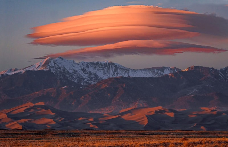 Stacked lens-shaped lenticular clouds lit orange by low sun above a snow-capped range, with a field of sand dunes across the middle of the frame