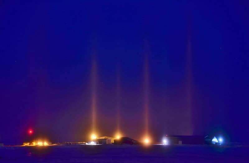 A row of vivid coloured vertical columns of light rising from ground lights along a horizon, against a deep blue night sky