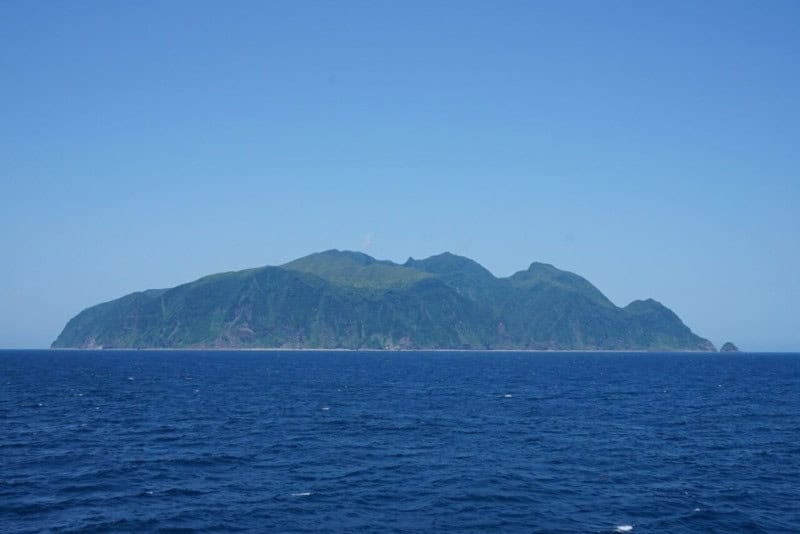 The steep green island of Mikura-jima seen from the sea under a clear blue sky, its summit hidden in a low cloud