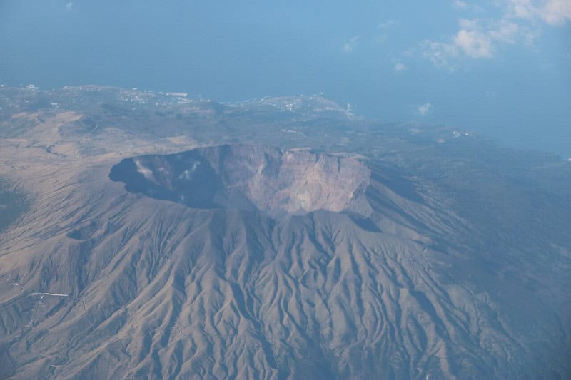 Aerial view of the wide grey summit crater of Mount Oyama on Miyake-jima, with ash-covered slopes and the coast beyond