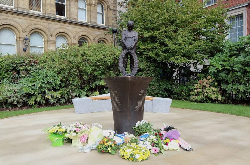 The MV Derbyshire memorial in Liverpool: a bronze figure on a plinth with bouquets of flowers laid at its base in a church garden