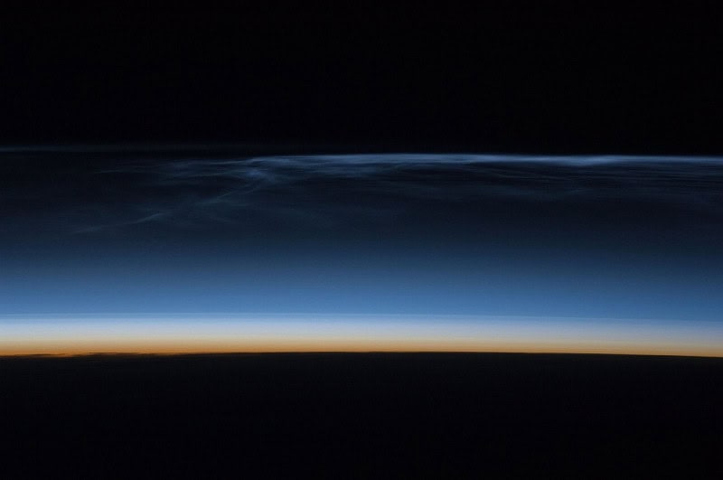 A thin bright band of noctilucent cloud glowing above the dark curve of the Earth in deep twilight, seen from orbit